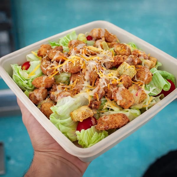 Hand holding a square container of salad with fried chicken, lettuce, tomatoes, cheese, and dressing.